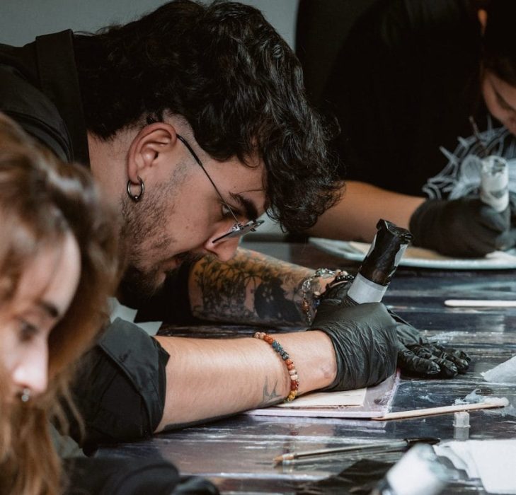Las personas trabajan intensamente en tareas detalladas, utilizando herramientas o dispositivos portátiles y usando guantes negros. Una persona en el foco tiene tatuajes en el brazo y está inclinada hacia adelante, concentrada en su trabajo. El entorno se asemeja a un taller o estudio donde podría estar teniendo lugar un curso de higiene sanitaria.