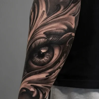 Tatuaje hiperrealista de un ojo con ornamentos en negro y gris en el brazo realizado en Avantgarde Tattoo Barcelona